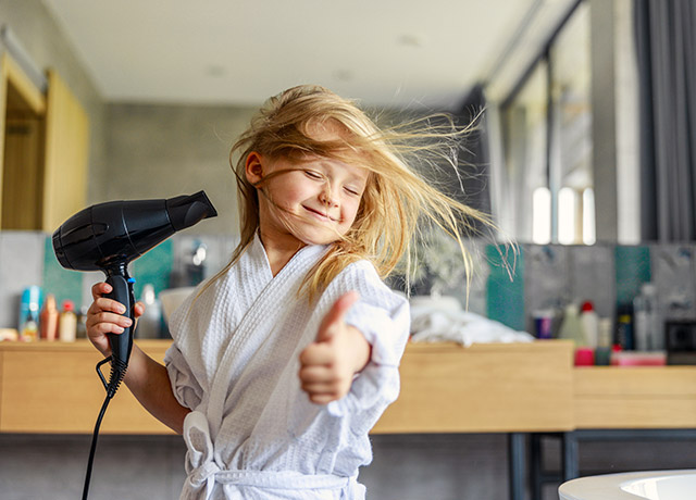 Kleines Mädchen zeigt Daumen hoch während sie sich die Haare föhnt